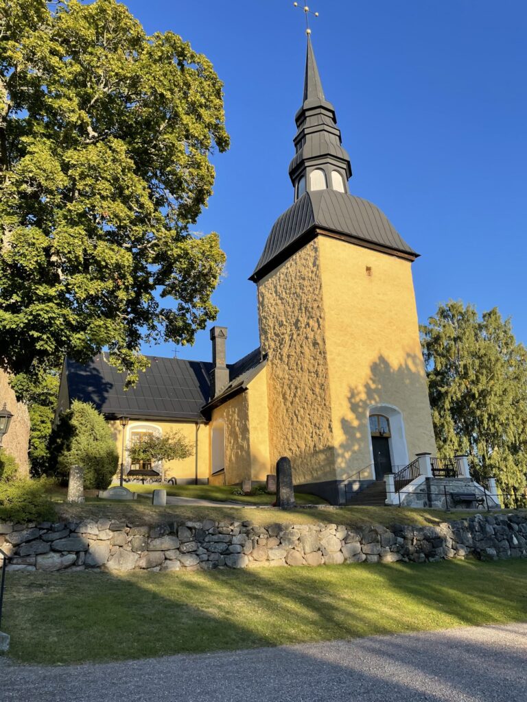 En gul kyrka med ett högt mörkt torn står mot en klarblå himmel. Solljus framhäver dess fasad. Ett stort träd skuggar kyrkogården som kantas av en stenmur och gräs. Skuggor av träden och strukturen är synliga på väggen.