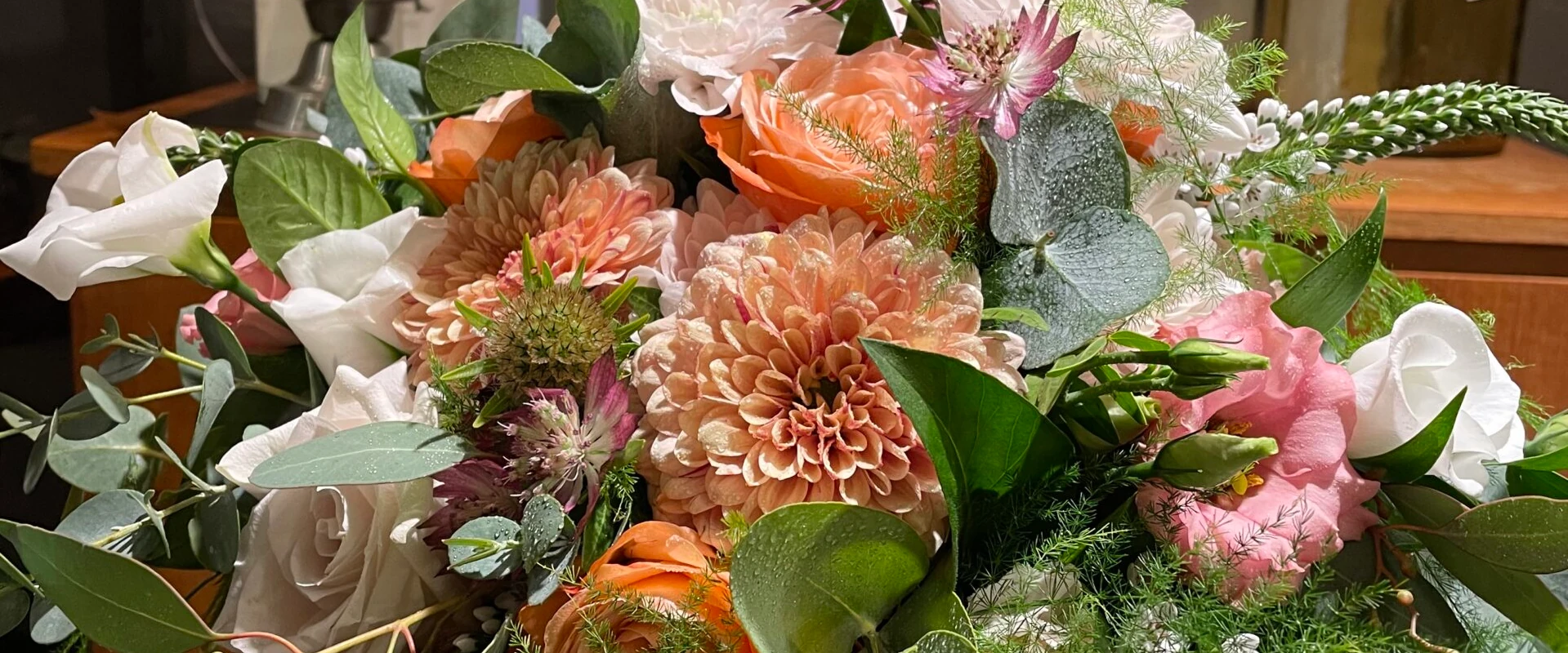 En livlig bukett med ett sortiment av blommor, inklusive orange dahlior, rosa rosor och frodig grönska. Arrangemanget är fylligt och färgglatt, vilket skapar en glad och elegant visning.