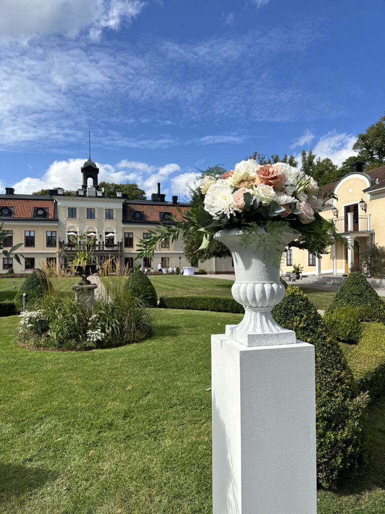 En vackert anlagd trädgård utanför en storslagen byggnad med en hög, vit pelare som stödjer en dekorativ vas fylld med ett arrangemang av rosa och vita blommor. Himlen är blå med spridda moln ovanför.