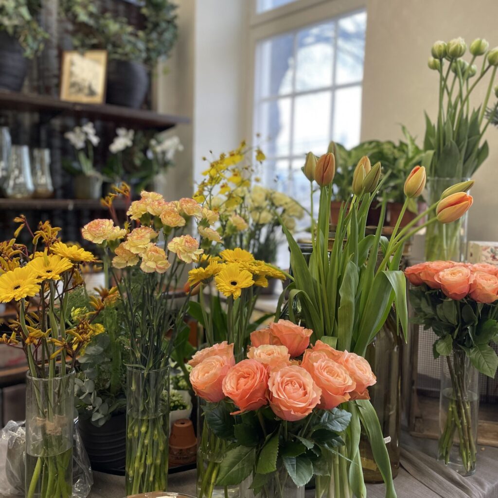 En mängd olika blommor i en butik, inklusive rosa rosor, gula prästkragar och ljusorange tulpaner, utställda i glasvaser på ett bord. Bakgrunden har ett stort fönster och ytterligare vaser på hyllor med dekorativa växter.