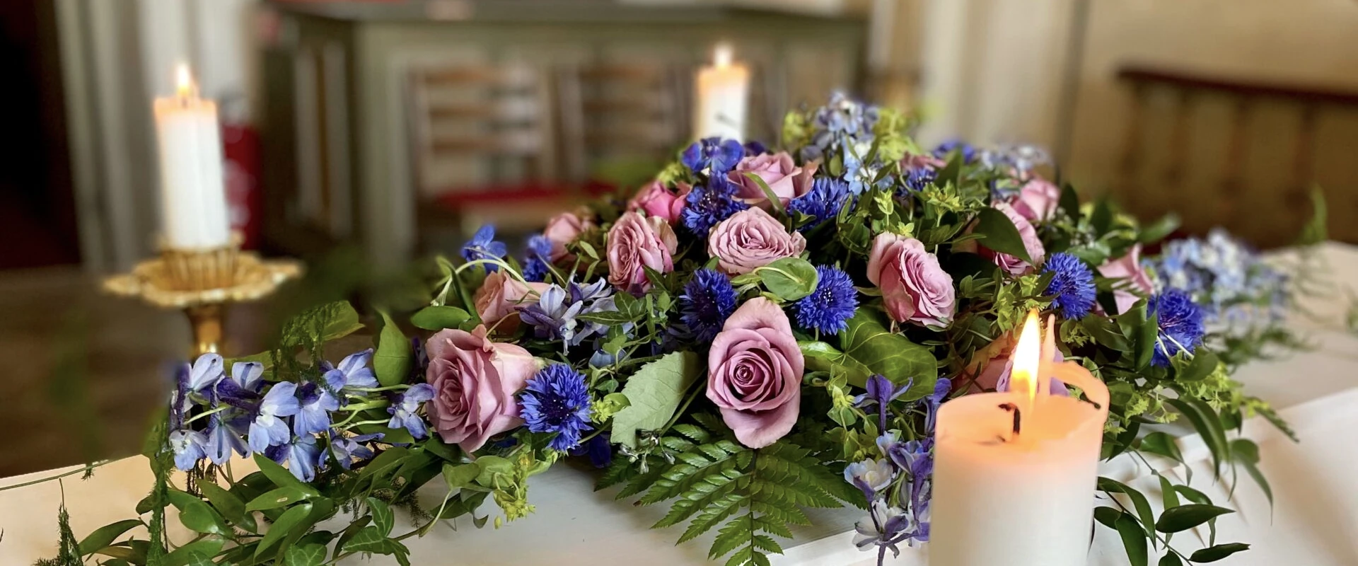 Ett blomsterarrangemang med lila rosor, blå blommor och frodiga gröna löv visas på ett bord. Tända vita ljus omger arrangemanget, vilket ger en varm glöd. Bakgrunden visar suddiga möbler och ett mjukt upplyst rum.