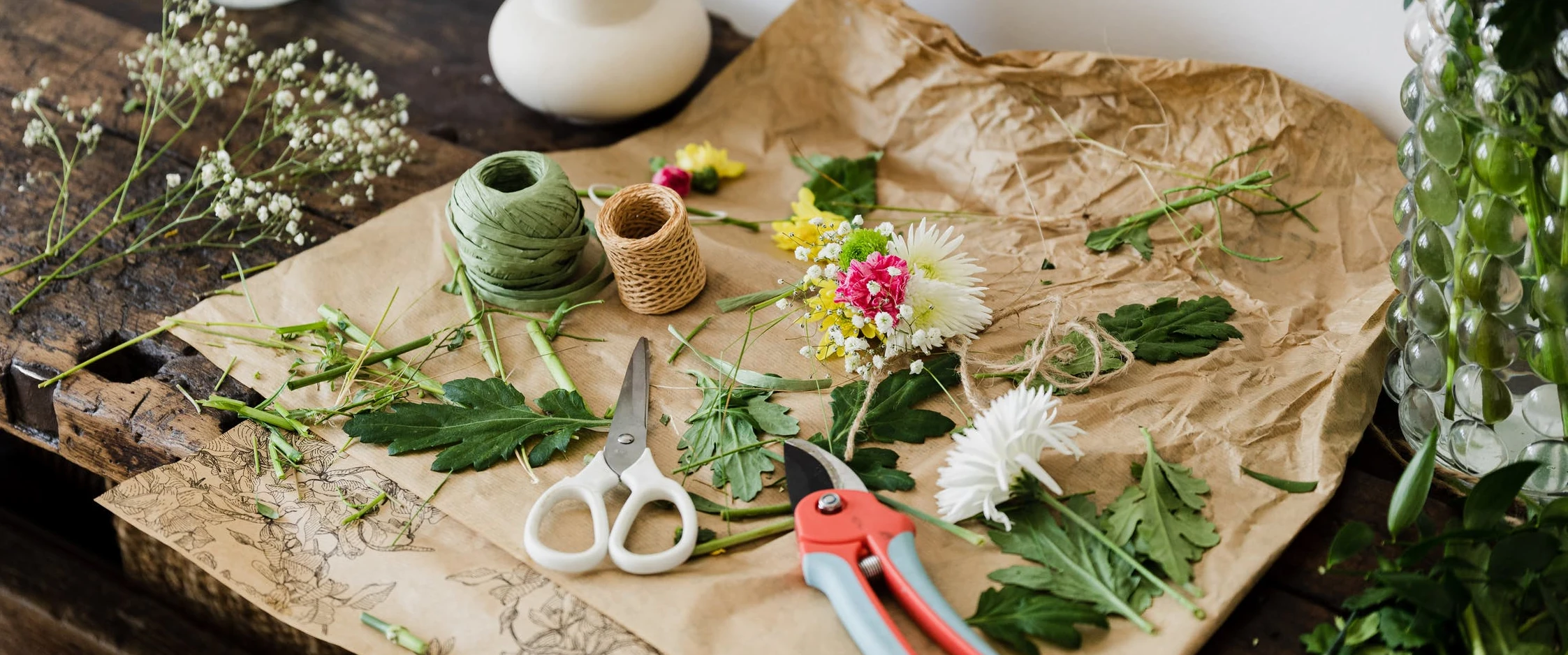 En rustik hantverksuppställning på ett träbord har brunt papper, nyklippta blommor, gröna blad, garn, beskärningssax och vit sax. Delika blommorna inkluderar små blommor och krysantemum, redo att arrangeras.