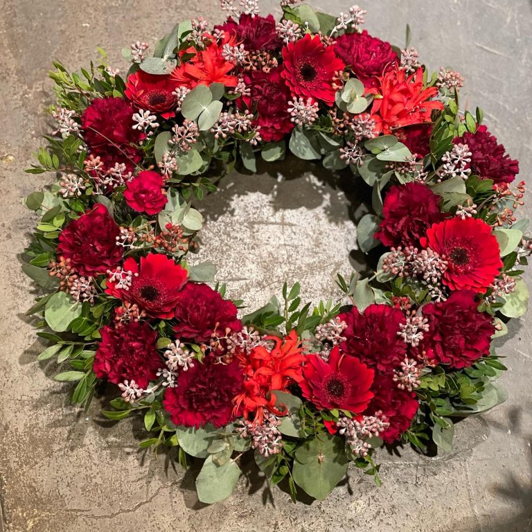 En cirkulär blomkrans med livfulla röda blommor, inklusive gerbera-prästkragar och nejlikor, accentuerad av gröna blad och små vita knoppar, placerade på en grov grå yta.