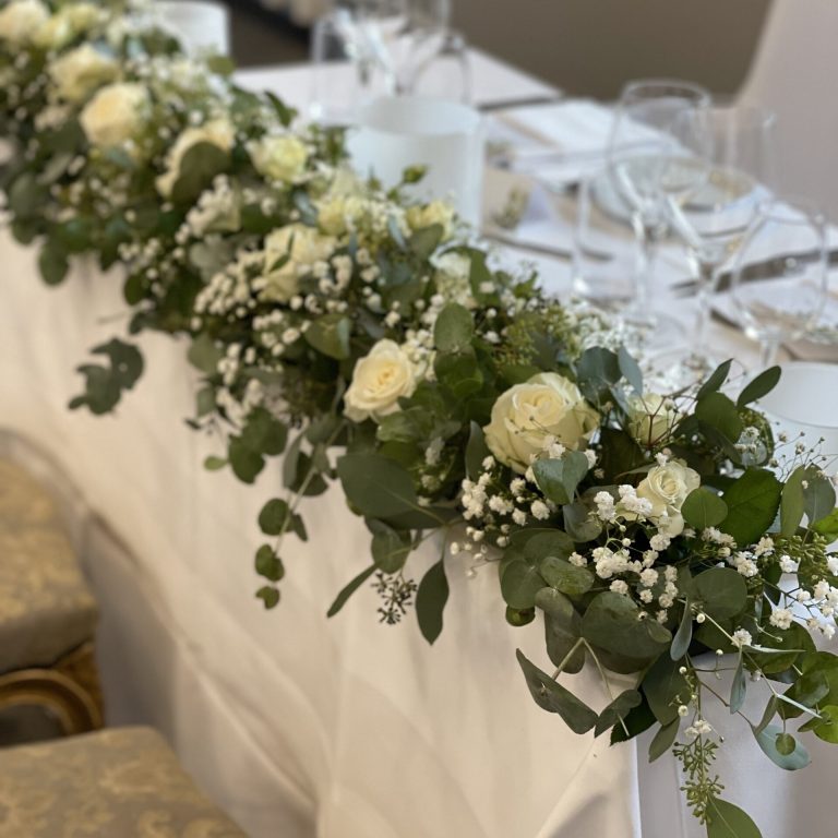 Ett bord prydt med ett elegant blomsterarrangemang med vita rosor, babys andedräkt och frodig grönska. Inramningen inkluderar vita dukar, kristallglas och prydligt arrangerade servetter, vilket skapar en sofistikerad restaurangatmosfär.