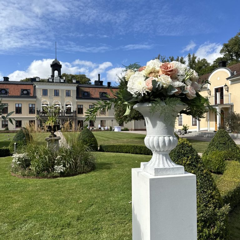 En vackert anlagd trädgård utanför en storslagen byggnad med en hög, vit pelare som stödjer en dekorativ vas fylld med ett arrangemang av rosa och vita blommor. Himlen är blå med spridda moln ovanför.